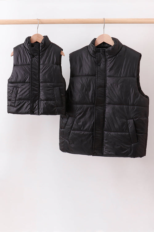 Two black puffer vests hanging on a rack against a white background